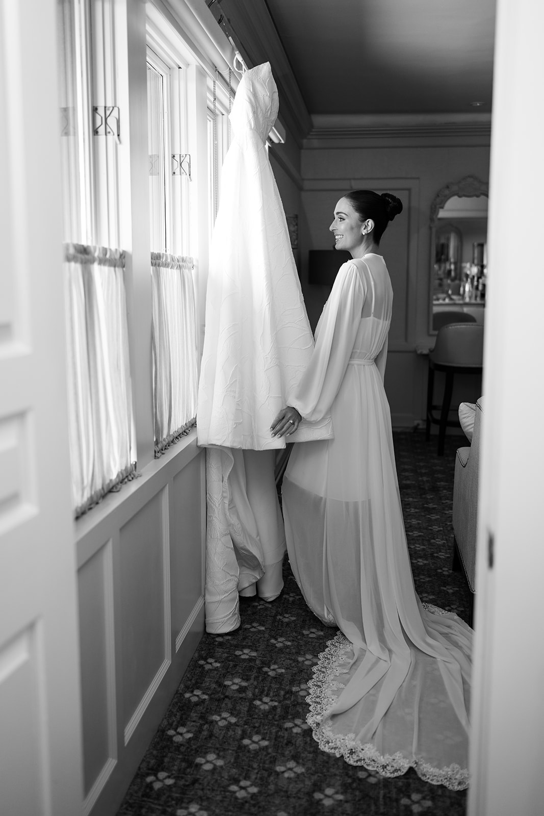 Black and white film portrait of a bride in a long robe, smiling as she looks at her hanging wedding dress.